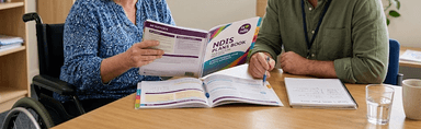 A support participant and a CDC team member sitting together at a table, going through an NDIS plan booklet page by page.
